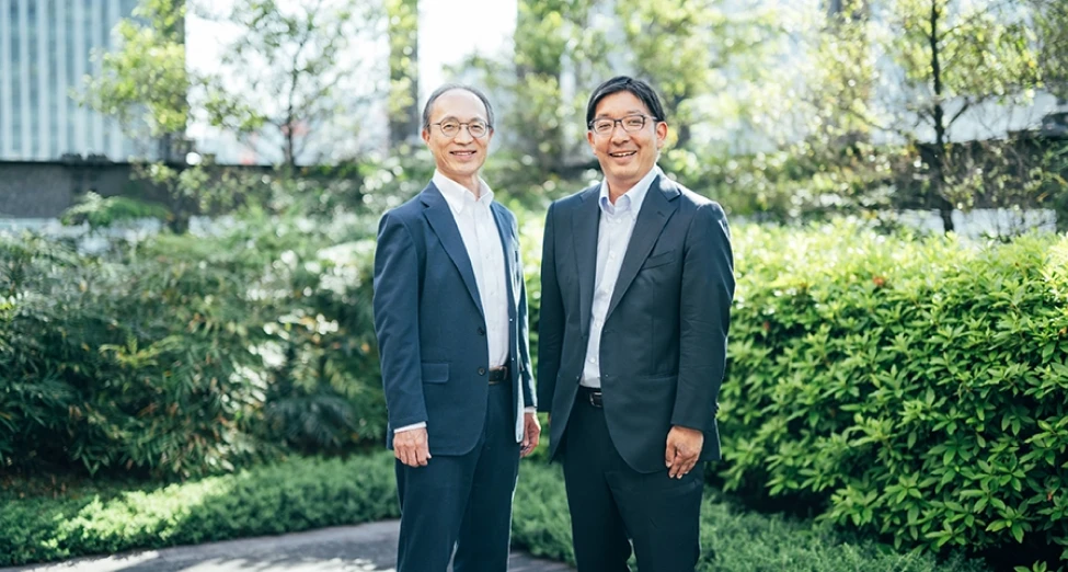 “Tomoyuki Tanaka, Senior Executive Officer and Group CSO, and Keisuke Miyake, Head of Corporate Communications, standing outdoors in front of greenery.”